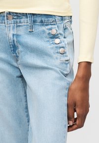 Ανοιχτό μπλε τζιν παντελόνι με τρία ορατά ασημένια κουμπιά στην περιοχή της μπροστινής τσέπης, συνδυασμένο με ένα κρεμ ριμπ μπλούζα μακρυμάνικη.