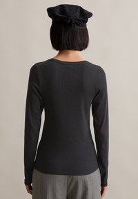Langärmliges, eng anliegendes schwarzes Oberteil mit geripptem Textur und Rundhalsausschnitt, kombiniert mit einem schwarzen Barett. Der Hintergrund ist neutral.