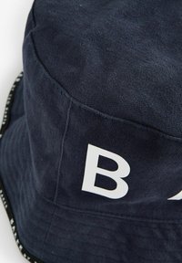 Pălărie de tip bucket din bumbac bleumarin, cu o literă albă „B” proeminentă pe lateral, având un material texturat și detalii cusute.