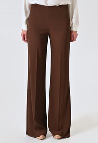 Pantaloni a gamba larga marroni realizzati in un tessuto morbido e liscio. Caratterizzati da un vita alta e un taglio lungo fino a terra, abbinati a calzature di colore chiaro.