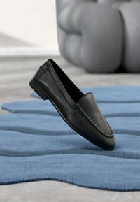 Chaussure slip-on en cuir noir à bout pointu et texture lisse, posée sur un tapis bleu ondulé.