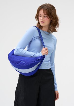 Frau trägt ein hellblaues Langarmshirt und schwarze Hose, sie trägt eine große blaue Schultertasche mit reflektierendem Streifen.