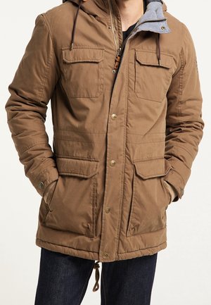 Homme portant une veste à capuche marron avec fermeture éclair et boutons, quatre poches avant à rabat et les mains dans les poches latérales.
