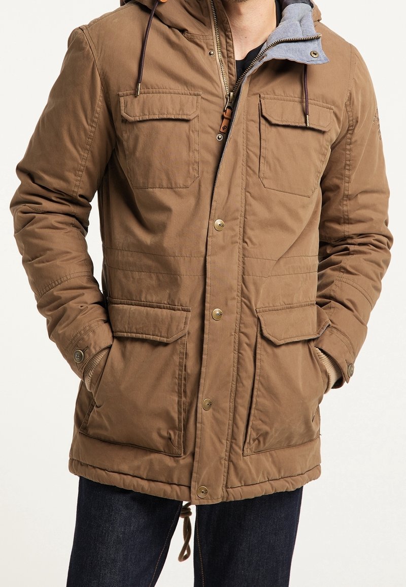 Homme portant une veste à capuche marron avec fermeture éclair et boutons, quatre poches avant à rabat et les mains dans les poches latérales.