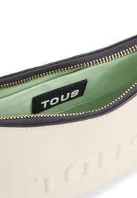 Kremowa saszetka z teksturowym wykończeniem i złotym zamkiem; wnętrze ma jasnozielone podszycie i czarną etykietę z logo.