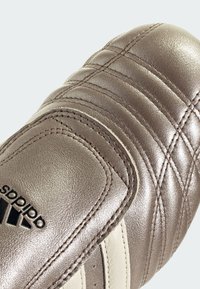 Metallic bronze soccer cleat with quilted texture, featuring three white stripes and black logo. Close-up view of the upper section.