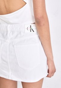 Biele denim mini sukňa s klasickým strihom a dvoma zadnými vreckami, ktorá má značkovú etiketu s nápisom "Calvin Klein Jeans" v mnohobarevných písmenách.