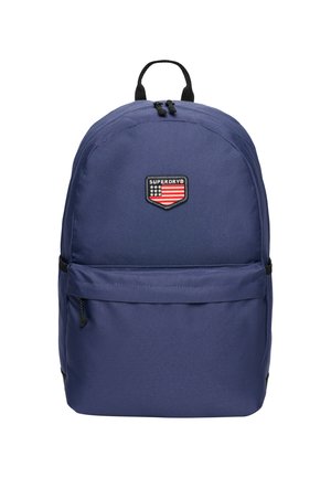 Mochila azul marino con un tejido duradero. Presenta un bolsillo frontal, cremalleras dobles y un parche de logo negro con un diseño de bandera.