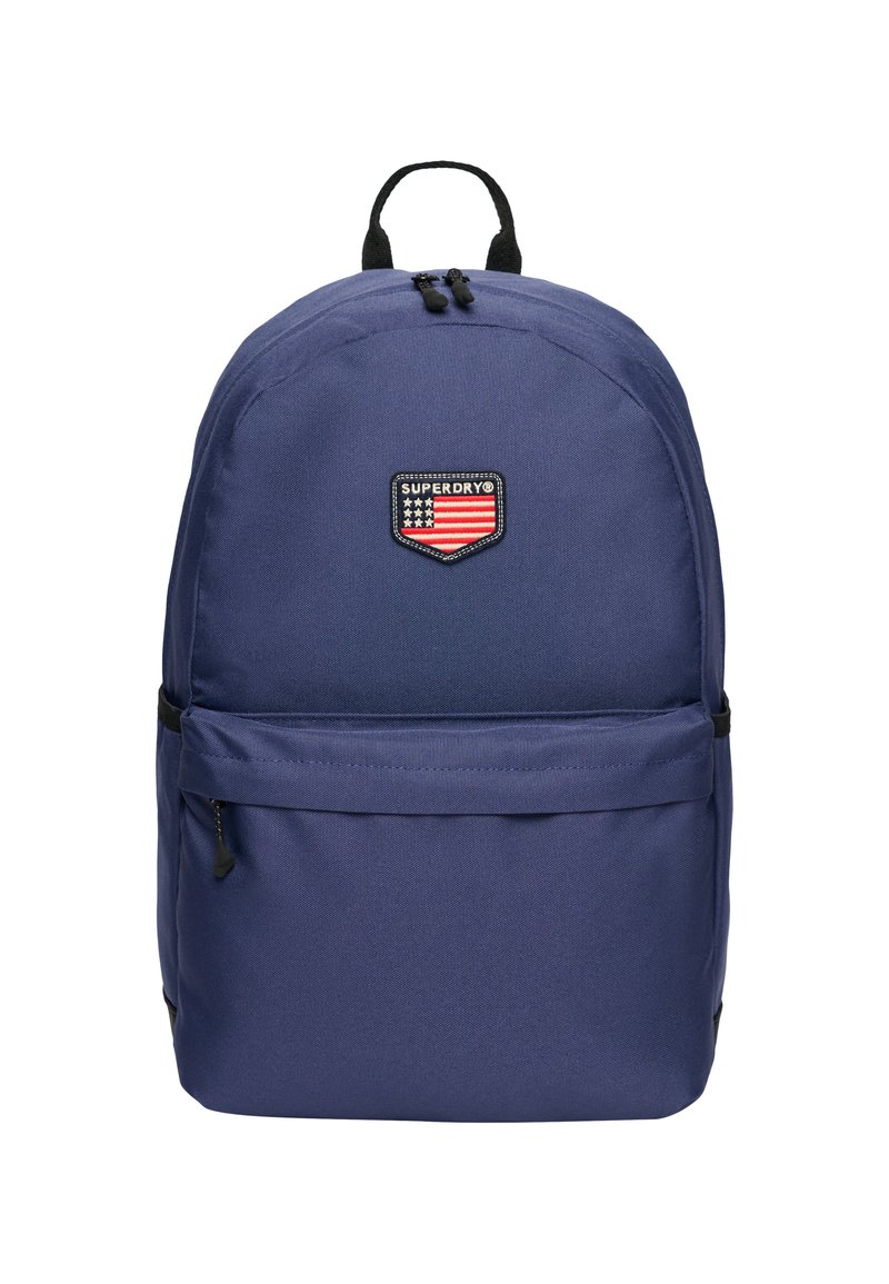 Mochila azul marino con un tejido duradero. Presenta un bolsillo frontal, cremalleras dobles y un parche de logo negro con un diseño de bandera.