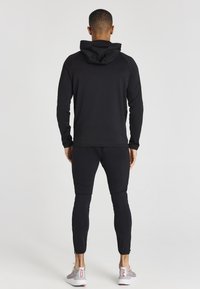 Černá mikina s kapucí na stažení, v kombinaci s dobře padnoucími černými tepláky. Hladká tkanina, bezešvý design a ribbované manžety. Sportovní boty s vzorovanými detaily.