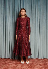 Femeie îmbrăcată într-o rochie midi florală roșu închis cu mâneci lungi și umeri bufanți, purtând pantofi albi ascuțiți, în fața unui fundal cu perdea albastră.