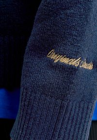 Suéter de punto azul marino oscuro con textura acanalada, que presenta texto dorado bordado "Originals Clothing" en la manga.