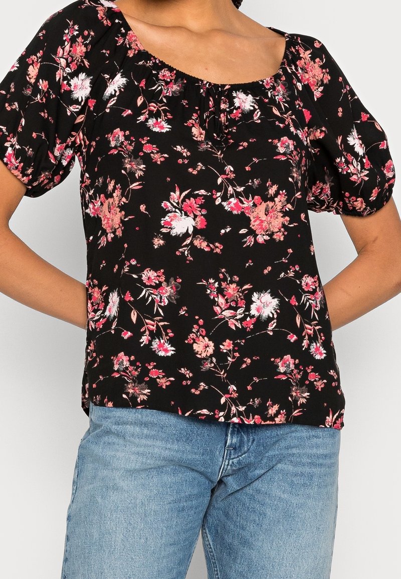 Blouse noire à manches courtes avec motif floral rose et blanc, manches bouffantes, associée à un jean bleu clair, mains du mannequin reposant dans le dos.