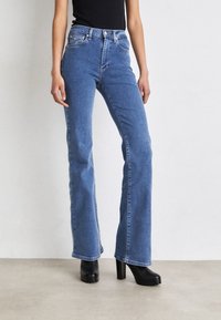 Woman wearing high-waist blue bootcut jeans and black heeled ankle boots standing against a plain background.