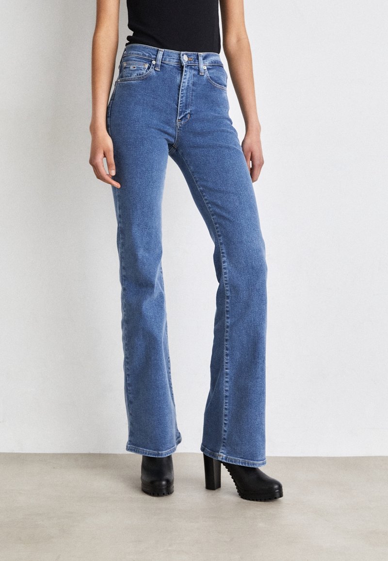 Woman wearing high-waist blue bootcut jeans and black heeled ankle boots standing against a plain background.