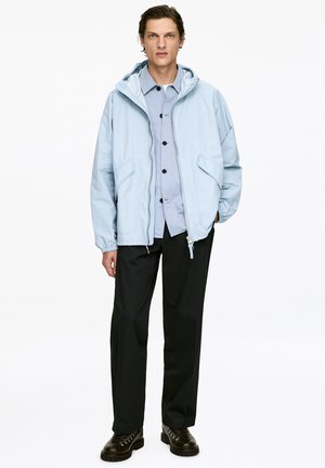 Light blue hooded jacket with a relaxed fit, two front pockets, worn over a button-up shirt, paired with black trousers and dark shoes.