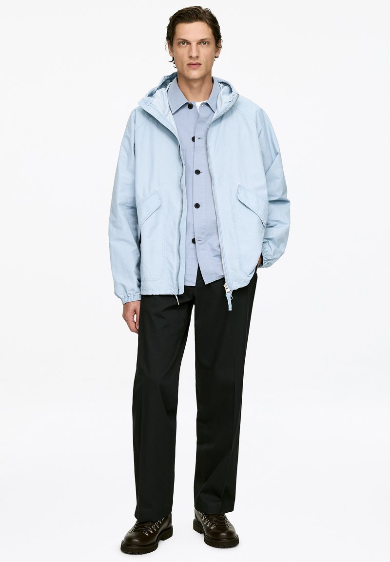 Veste à capuche bleu clair à coupe décontractée, avec deux poches avant, portée sur une chemise boutonnée, associée à un pantalon noir et des chaussures foncées.