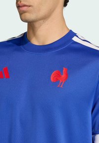 Modré sportovní tričko z hladké látky s červeným logem kohouta a červenými pruhy Adidas na ramenou. Kulatý výstřih.