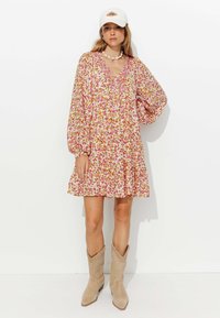 Robe florale à manches longues, motifs roses et jaunes, encolure en V, taille froncée, associée à des bottes de cowboy beiges et à une casquette blanche.