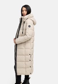 Cappotto imbottito lungo beige con cappuccio, caratterizzato da una texture trapuntata, cerniera scura, tasche laterali e un patch circolare con logo sulla manica.