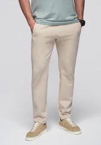 Man in beige slim-fit broek, beige sneakers met witte veters, en een lichtgrijs T-shirt, staande met handen in de zakken.