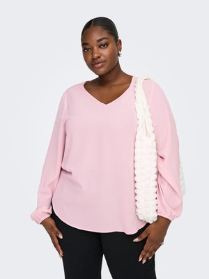 Młoda kobieta ubrana w różową bluzkę z długim rękawem i czarne spodnie, nosząca na prawym ramieniu teksturowaną białą torbę na ramię.