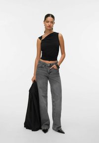 Top nero senza maniche con texture arricciata, design asimmetrico e dettagli in oro; abbinato a jeans grigi a gamba larga e scarpe nere con punta a stiletto.