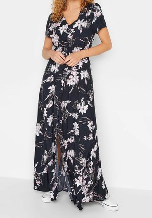 Vrouw draagt een zwarte maxi-jurk met bloemenprint, korte mouwen en knopen aan de voorkant, gecombineerd met witte sneakers.