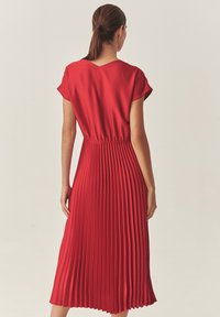 Červené skladané šaty s krátkymi rukávmi, zúženým pásom a splývavou sukňou. Hladká textília so štruktúrovaným dizajnom skladov zvyšuje pohyb.