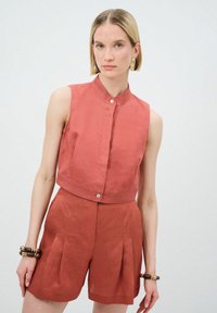 Rustfarbene ärmellose Bluse mit hohem Kragen und vorderer Knopfverschluss, aus einem strukturierten Stoff gefertigt. Kombiniert mit passenden Shorts mit Falten.