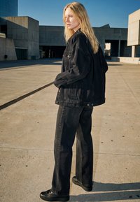 Lyshåret person i sort denimjakke og bukser står på en betonplads nær moderne bygninger under klar blå himmel.