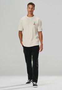 Beige T-Shirt mit kurzen Ärmeln und grafischem Design auf der Brust, kombiniert mit schwarzen geraden Hosen und schwarzen Sneakers. Einfacher, lässiger Look.