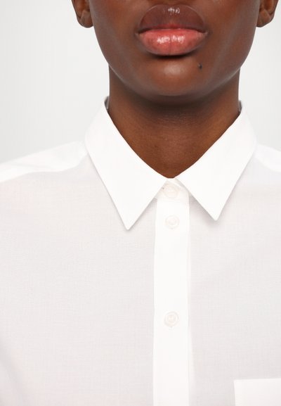 Camicia bianca con colletto, dal tessuto morbido e con taschino frontale nascosto. Presenta bottoni madreperlacei e un design essenziale.