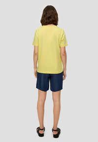 T-shirt en coton jaune à manches courtes, associé à un short en denim foncé. Sandales noires avec des lanières tissées. Fond neutre.