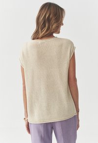 Beige pletený svetr s krátkými rukávy, kulatým výstřihem a texturovaným materiálem, v kombinaci se světle fialovými kalhotami.