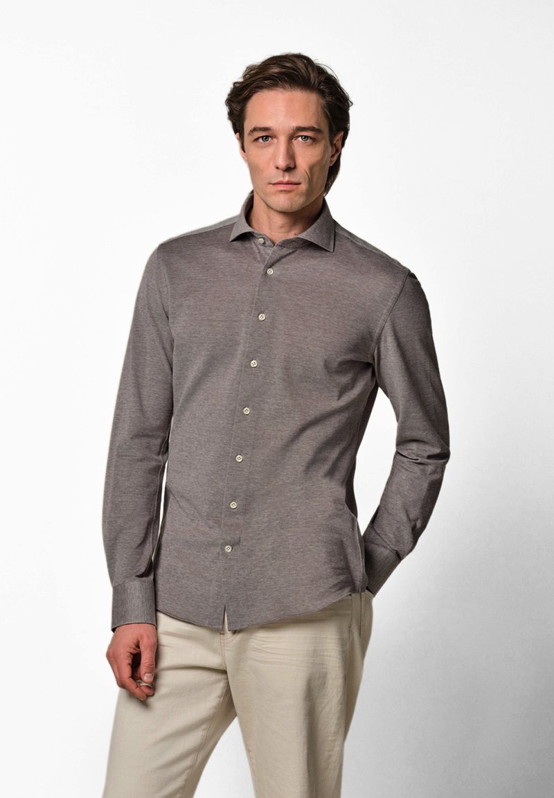 Hombre de pie mirando hacia adelante, vestido con una camisa gris ajustada abotonada y pantalones beige claros, sobre un fondo blanco liso.