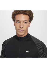 Schwarze Sportjacke mit hohem Kragen, ausgestattet mit Streifenmuster auf den Schultern und einem weißen Nike-Logo auf der Brust. Glatte Textur.