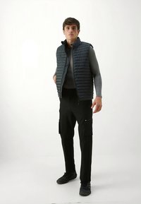 Vadderad marinblå väst över en grå tröja, kombinerad med svarta cargobyxor. Märkbara detaljer är sidofickor och dragkedja. Svarta sneakers.