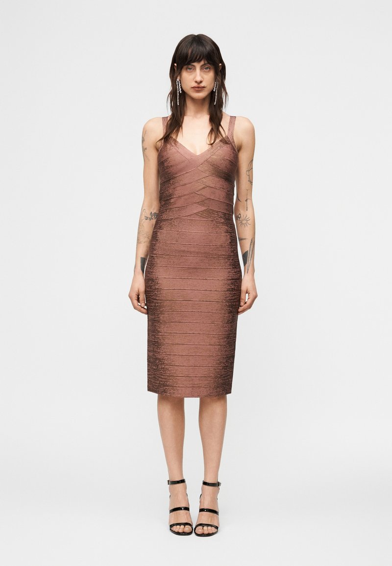 Γυναίκα με εφαρμοστό, αμάνικο, υφασμάτινο φόρεμα μήκους μέχρι το γόνατο σε χρώμα ρόδου καφέ και μαύρα ψηλοτάκουνα με λουράκι στον αστράγαλο, που στέκεται μπροστά σε λευκό φόντο.