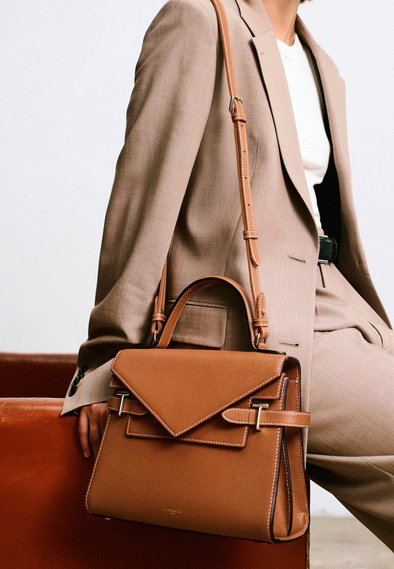 Borsa a tracolla in pelle marrone con un design strutturato, una pattina triangolare, accessori in metallo dorato e cuciture a contrasto. Include una tracolla regolabile.