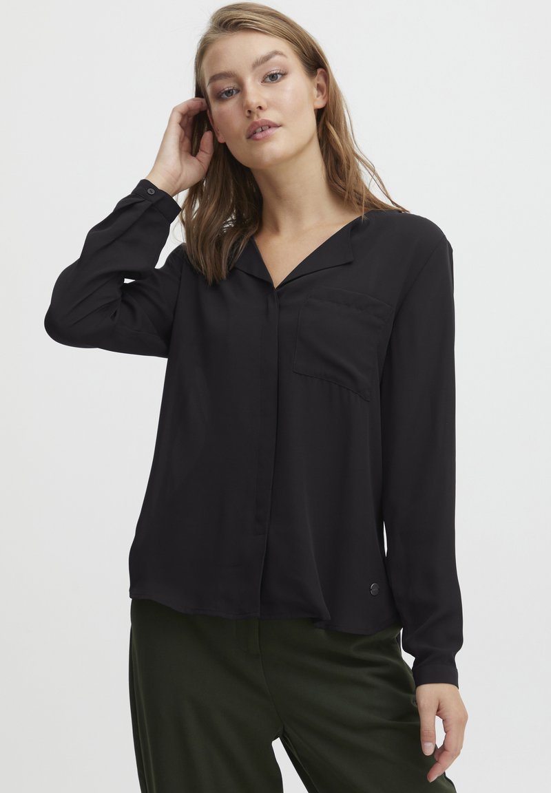 Blouse noire avec col, manches longues, poche poitrine unique et coupe légèrement décontractée; fabriquée en tissu lisse avec un léger éclat.