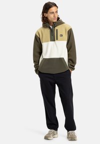 Mikina s kapucí z fleecu s barevným blokovým designem: béžová, olivově zelená a bílá. Obsahuje polo-zip, nastavitelnou kapuci a přední klokaní kapsu.