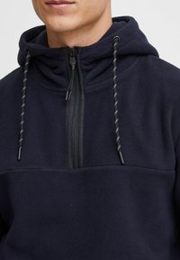 Donker marineblauwe fleece pullover met een ritskraag, voorzien van een capuchon en trekkoorden met een zwart en geel patroon. Zachte textuur en eenvoudig ontwerp.