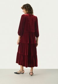Lång burgundyröd sammetsklänning med utsvängda ärmar, lagerdesign och en flödande silhuett, i kombination med svarta slingback-skor.