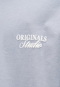 Ανοιχτό γκρι βαμβακερό μπλουζάκι με λευκό κεντημένο κείμενο "ORIGINALS Studio" σε casual καλλιγραφική γραμματοσειρά. Λείο υφή, απλός σχεδιασμός.