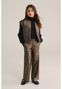 Metāliski bronzas divdaļīgs apģērbs, kas sastāv no bezpiepes žaketes un platām biksēm, apvienots ar melnu ribotā stila turtleneck un melnām kurpēm.