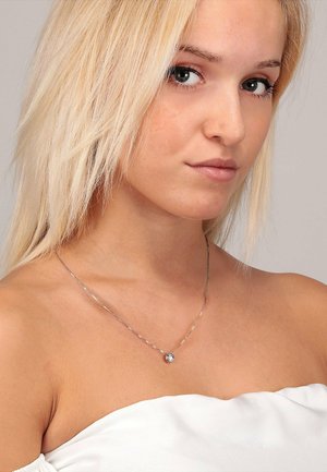 Silberne Halskette mit einer dünnen Kette und einem runden Anhänger; getragen auf nackter Haut und ergänzt ein schulterfreies weißes Oberteil.