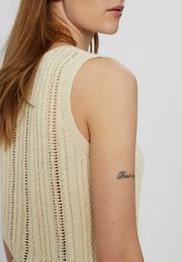 Beige gebreide mouwloze top met verticale ribstructuur en open stiksels op de rug. Zichtbare tattoo op de bovenarm.