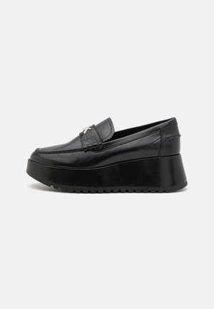 Mocasín de plataforma de cuero negro con suela gruesa y hebilla plateada decorativa en la parte superior, mostrado en perfil lateral sobre fondo blanco.