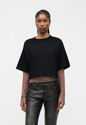 Frau mit geflochtenem Haar, die ein schwarzes, kurzes, bauchfreies Oberteil über einem langärmligen braunen Shirt und schwarzen, strukturierten Hosen trägt.